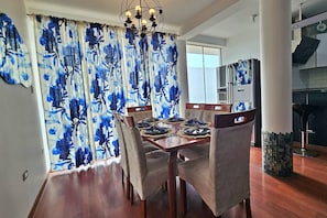 Uma sala de jantar com mesa de madeira posta para quatro pessoas, rodeada por cadeiras, e uma cozinha com geladeira e banquetas.