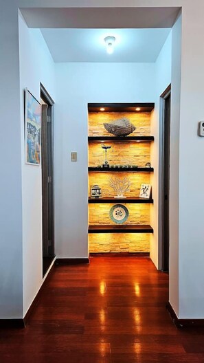 A hallway with wooden flooring, a wall-mounted shelf with decorative items, and a ceiling light.