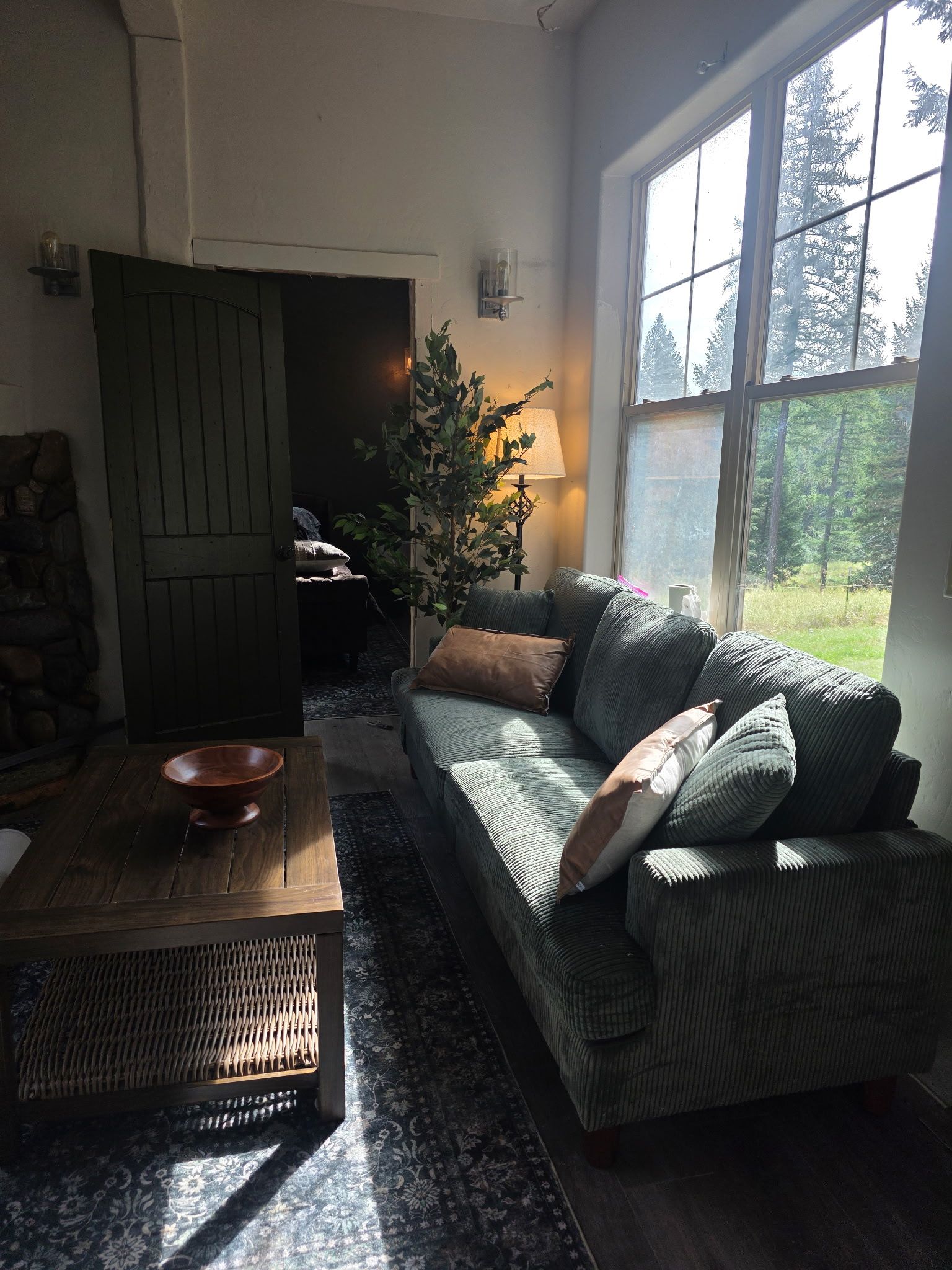 A cozy living room with a sofa, a coffee table, and a lamp. A door leads to another room, and there's a window with a view of trees.