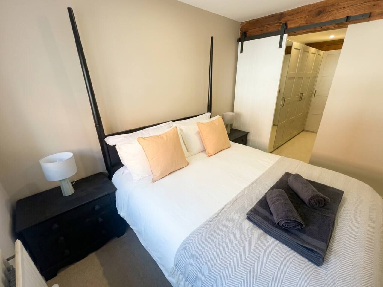 Una cama bien hecha con una colcha blanca y estampada, dos almohadas, una mesita de noche con una lámpara y una toalla enrollada sobre la cama.