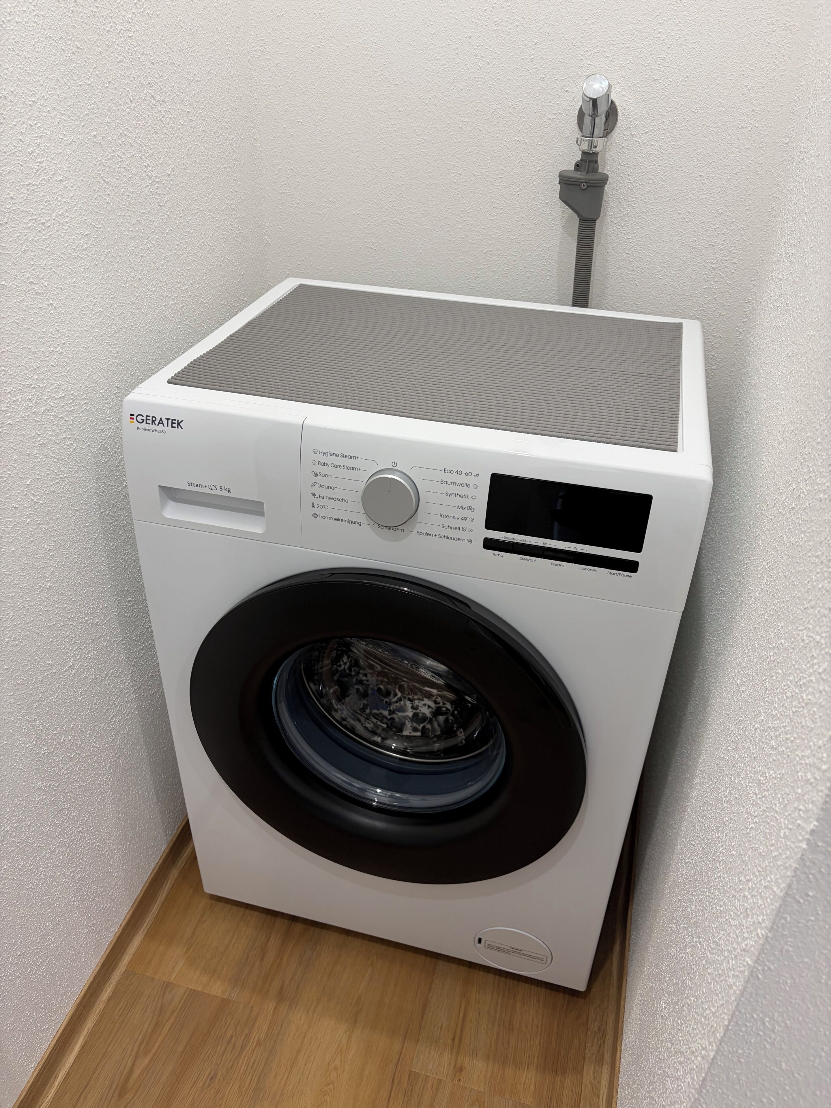 Eine weiße Waschmaschine mit Frontlader, einer Bedienfeld, einem Wasserschlauch und Holzboden.