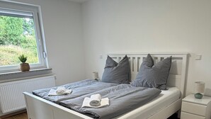 Quarto com cama, mesa de cabeceira, janela e vista para o verde lá fora.