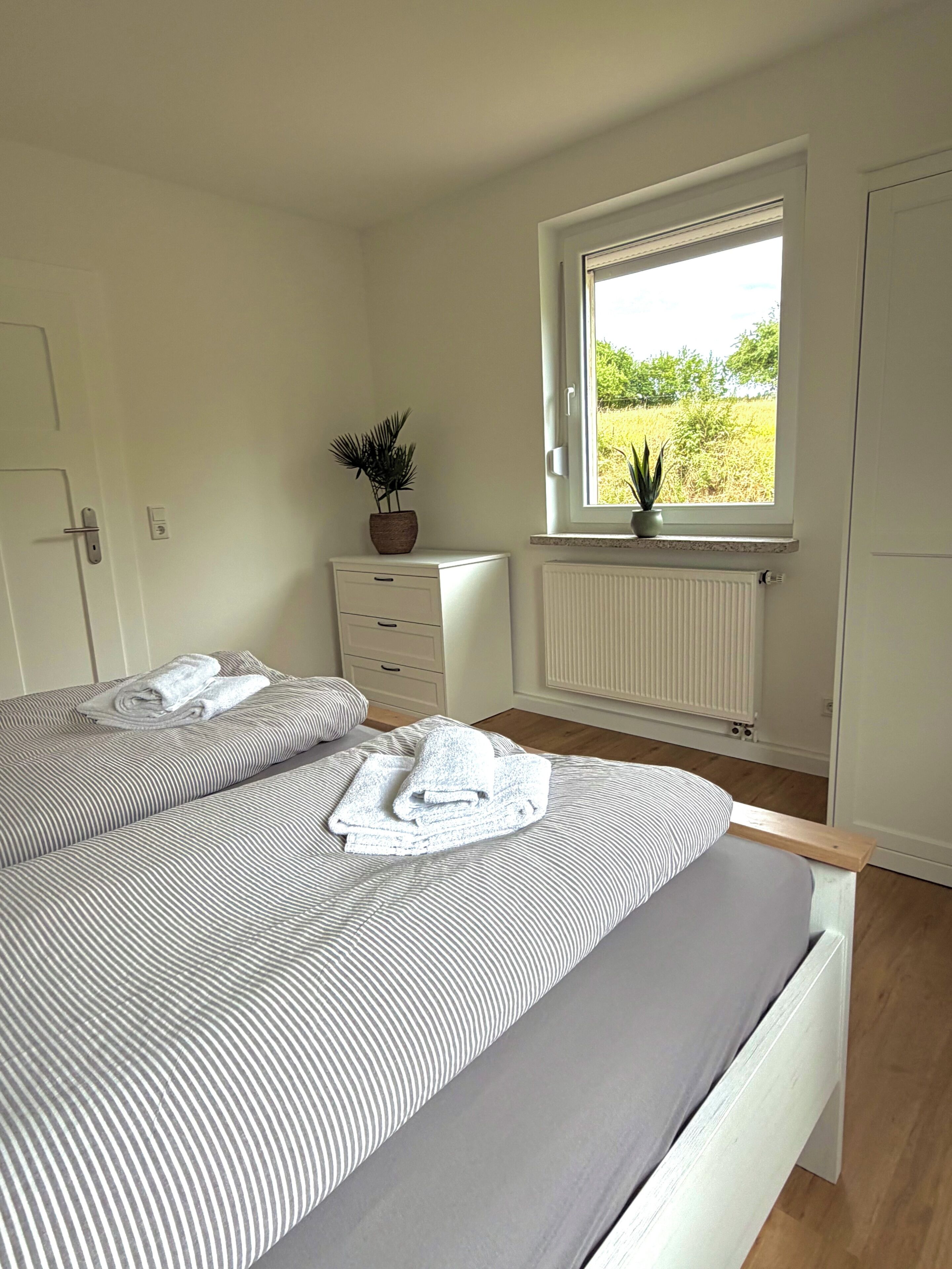 Une chambre avec deux lits, une fenêtre donnant sur un espace verdoyant, un radiateur et une plante en pot.
