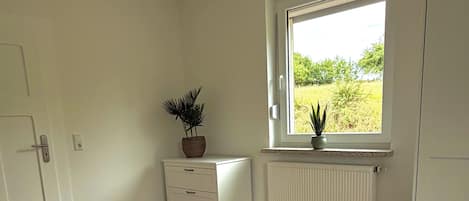 A bedroom with two beds, a window with a view of greenery, a radiator, and a potted plant.