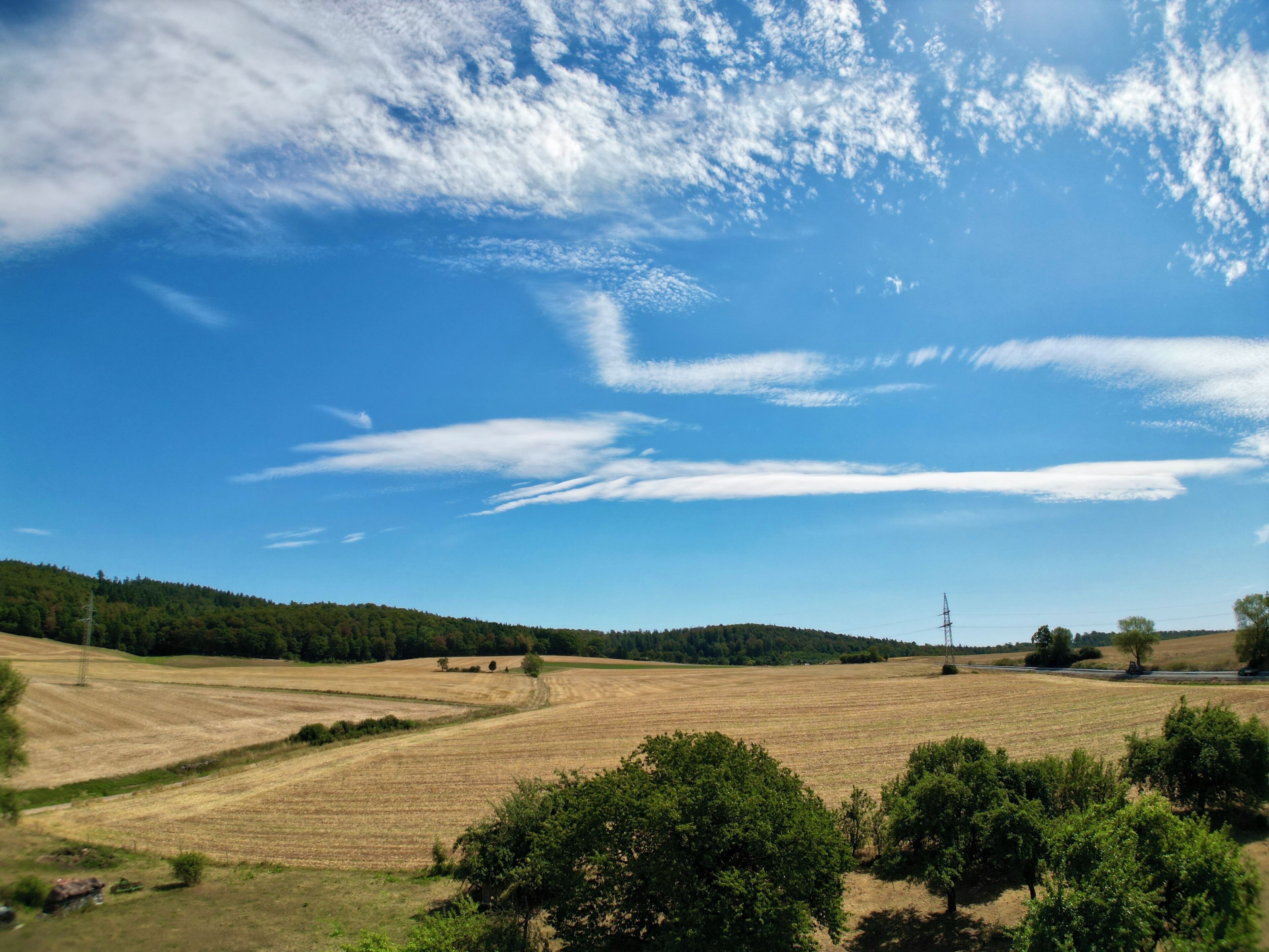 Ländliche Landschaft mit sanften Hügeln, Feldern und einem klaren, blauen Himmel mit vereinzelten Wolken.