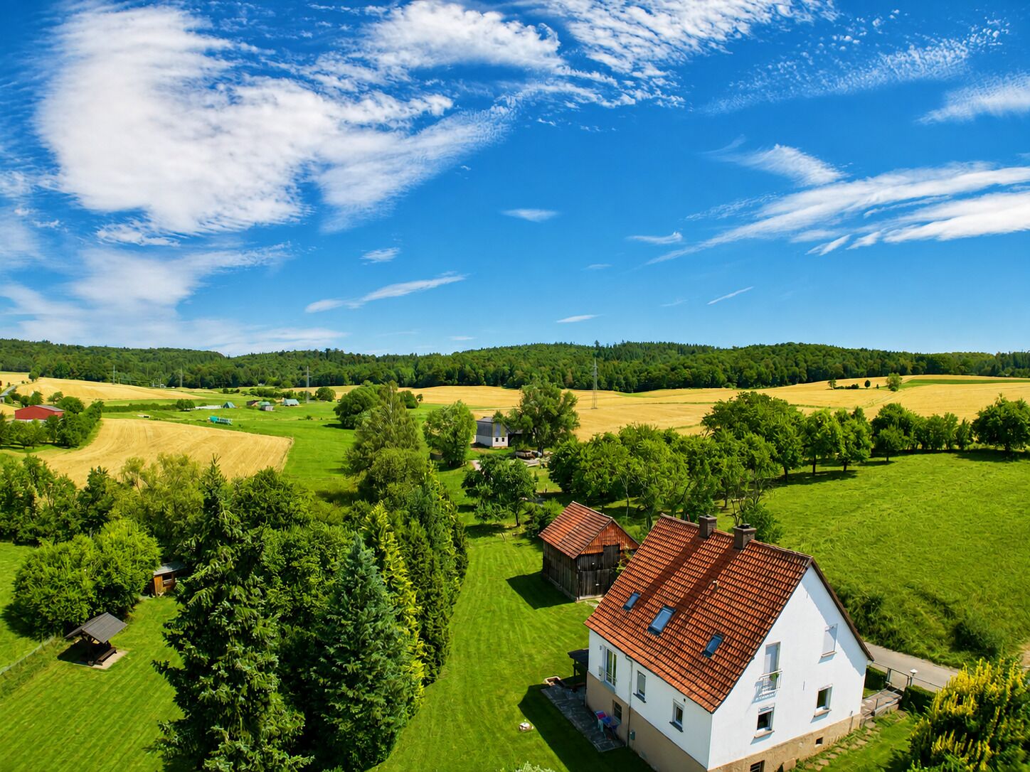 Landelijk landschap met een wit huis met een rood dak, omringd door groene velden en bomen.