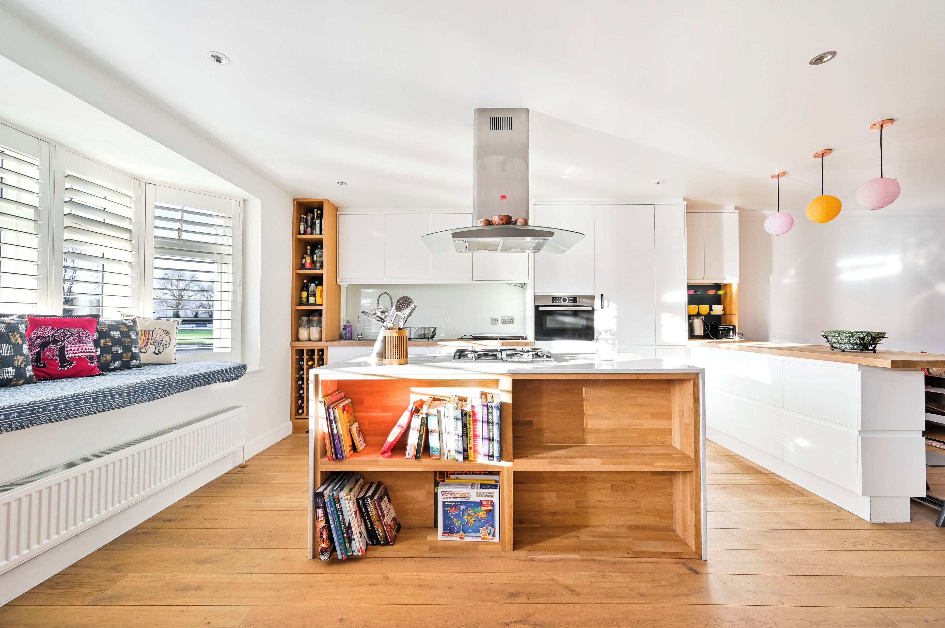 A modern kitchen with a central island, wooden shelves, and a window with blinds.