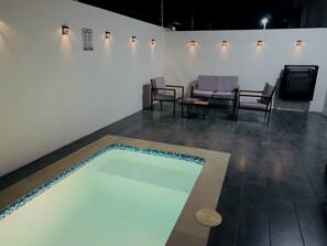 Zona de piscina amb vora enrajolada, saló amb sofà i cadires, i aplics murals.