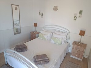 Ein ordentlich bezogenes Bett mit Kissen und gefalteten Handtüchern, ein Nachttisch mit Lampe sowie ein Spiegel an der Wand.