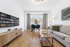 A modern living room with a flat-screen TV mounted on the wall, a wooden console table, a sofa, a dining area with chairs, and a large window with curtains. - Perfect Location near Heathrow & 2 min London Tube (Hounslow)