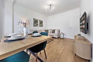 A modern living room with a dining table set for four, a sofa, a TV mounted on the wall, and a small side table. - Perfect Location near Heathrow & 2 min London Tube (Hounslow)