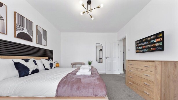 A bedroom with a bed, a wooden dresser, a wall-mounted TV, and a ceiling light fixture. - Perfect Location near Heathrow & 2 min London Tube (Hounslow)