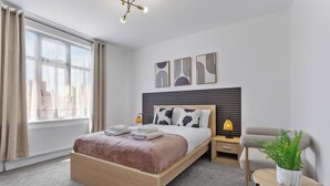 A modern bedroom with a wooden bed, a bedside table with a lamp, a small round table with a plant, and artwork on the wall. - Perfect Location near Heathrow & 2 min London Tube (Hounslow)