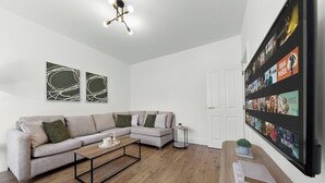 A modern living room with a sectional sofa, a coffee table, and a wall-mounted TV displaying a movie. - Perfect Location near Heathrow & 2 min London Tube (Hounslow)