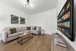 A modern living room with a sectional sofa, a coffee table, and a wall-mounted TV displaying a movie. - Perfect Location near Heathrow & 2 min London Tube (Hounslow)