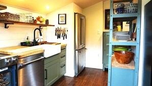 Eine Küche mit einem Edelstahl-Kühlschrank, einer Spüle und einem Herd. Messer hängen an der Wand und ein Regal mit Geschirr ist vorhanden.
