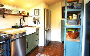 A kitchen with a stainless steel refrigerator, a sink, and a stove. There are knives hanging on the wall and a shelf with dishes. - Charming little cottage minutes from town (Gibsons)