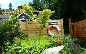 A wooden fence with a 'PARKING' sign, a lifebuoy, and a stone with the number '350' in a landscaped area. - Charming little cottage minutes from town (Gibsons)