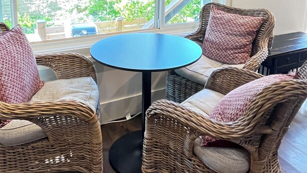 Ein Zimmer mit Korbmöbeln, einem runden Tisch und Blick durch große Fenster.