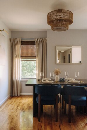 Uma sala de jantar com mesa de madeira posta para quatro pessoas, luminária pendente e janela com cortinas.