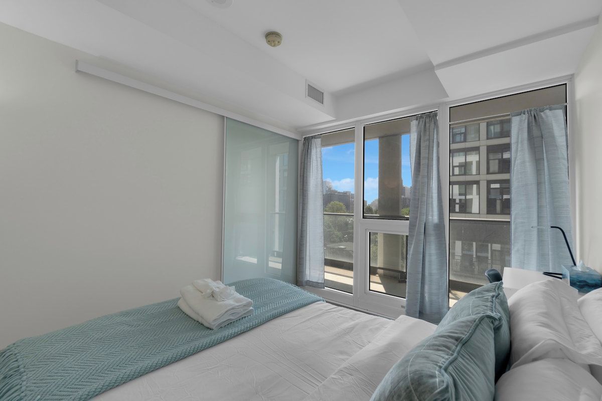 Hotellihuone, jossa on sänky, ikkuna verhoilla, näkymä kaupunkiin ja ovi, joka johtaa ulkotilalle.