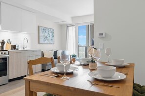 Una cocina moderna con armarios blancos, una mesa de comedor para dos personas con platos y copas de vino blancas, y vistas a la ciudad a través de un ventanal grande.