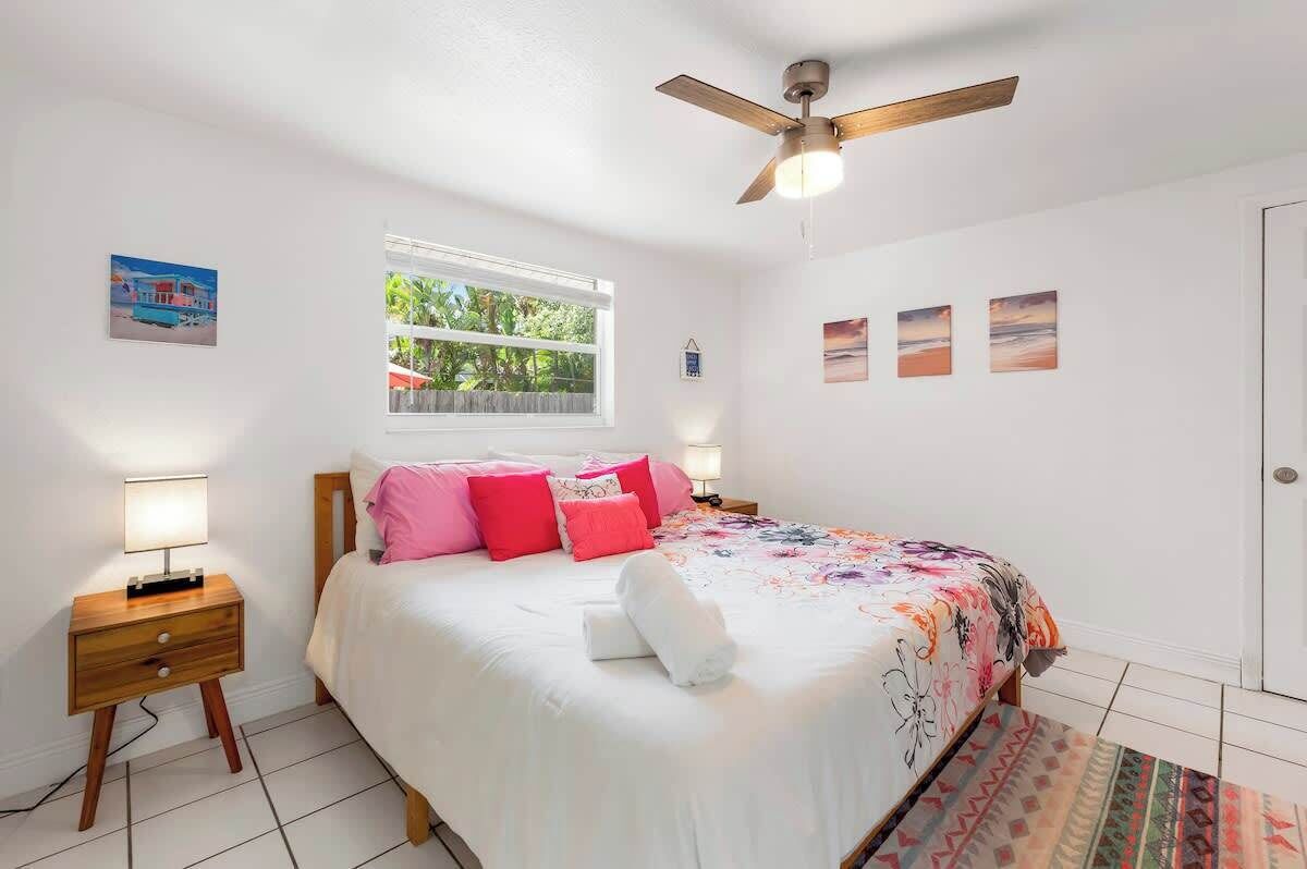 Un dormitorio con cama, mesita de noche, ventilador de techo, ventana y decoración en la pared.