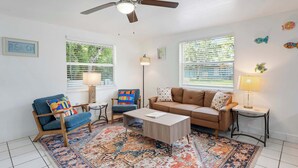 Ein Wohnzimmer mit Deckenventilator, zwei Fenstern mit Jalousien, einem Sofa, einem Sessel, einem Couchtisch und einem gemusterten Teppich.