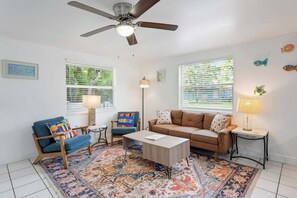 Ein Wohnzimmer mit Deckenventilator, zwei Fenstern mit Jalousien, einem Sofa, einem Sessel, einem Couchtisch und einem gemusterten Teppich.