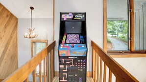 Uma escadaria com corrimãos de madeira, um candelabro e uma máquina de jogos de fliperama antiga com a inscrição 'Arcade Up' em um ambiente com janela.