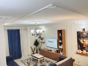 Un salon avec un téléviseur à écran plat, une table basse, un canapé, un rideau bleu et un lustre.