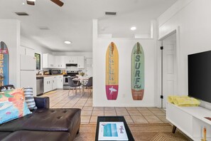 Ein modernes Wohnzimmer mit einem braunen Ledersofa, einem Couchtisch mit einer Zeitschrift und einem Fernseher auf einem weißen Schrank. An der Wand hängen Surfbretter, und im Hintergrund ist eine Küche zu sehen.