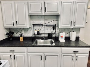A kitchen with white cabinets, a black countertop, a stainless steel sink, a coffee maker, and a paper towel holder.