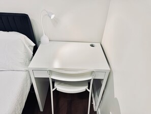 A small, white desk with a lamp, positioned next to a bed in a room with a dark wooden floor.