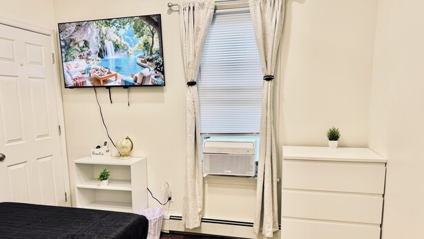 A bedroom with a bed, a TV mounted on the wall, a window with curtains, a small shelf with a globe and a plant, and a white dresser.