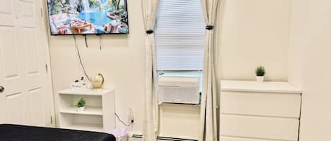 Un dormitorio con cama, televisor montado en la pared, ventana con cortinas, un estante pequeño con un globo terráqueo y una planta, y un aparador blanco.