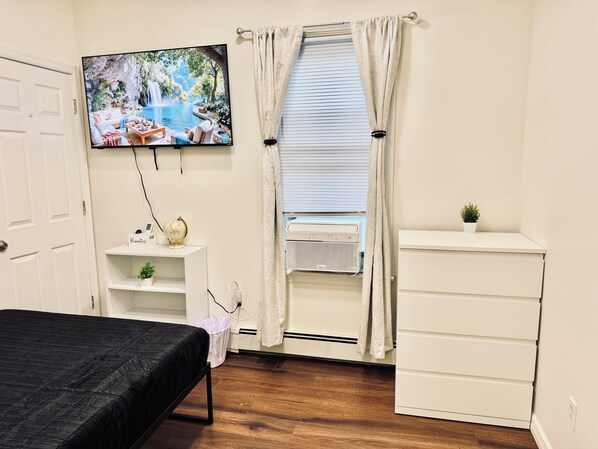 A bedroom with a bed, a TV mounted on the wall, a window with curtains, a small shelf with a globe and a plant, and a white dresser.