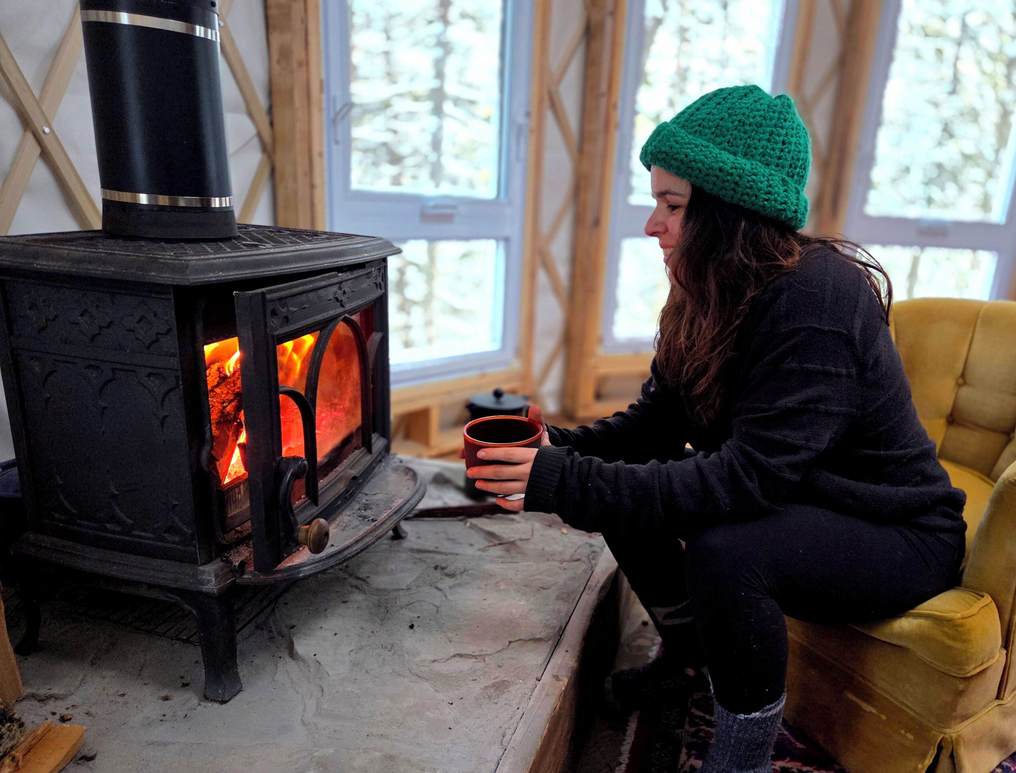 A person seated by a wood-burning stove, holding a mug.