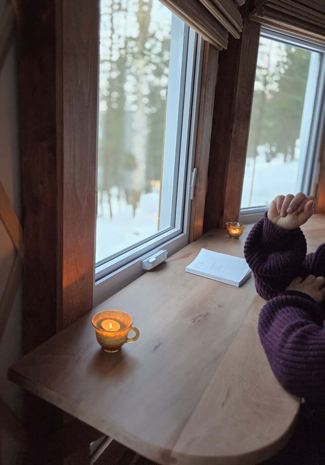 Une table en bois, une bougie, un carnet et une personne assise près d’une fenêtre, avec vue sur les arbres.