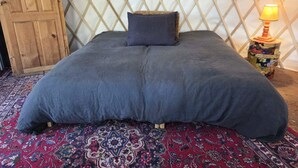 A bed with a dark blue bedspread and a single pillow, a patterned rug, a wooden door, and a small table with a lamp.