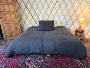A bed with a dark blue bedspread and a single pillow, a patterned rug, a wooden door, and a small table with a lamp.