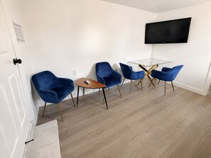 Ein modernes Wohnzimmer mit einem an der Wand befestigten Flachbildfernseher, einem Glastisch und vier blauen Stühlen mit goldenen Beinen.