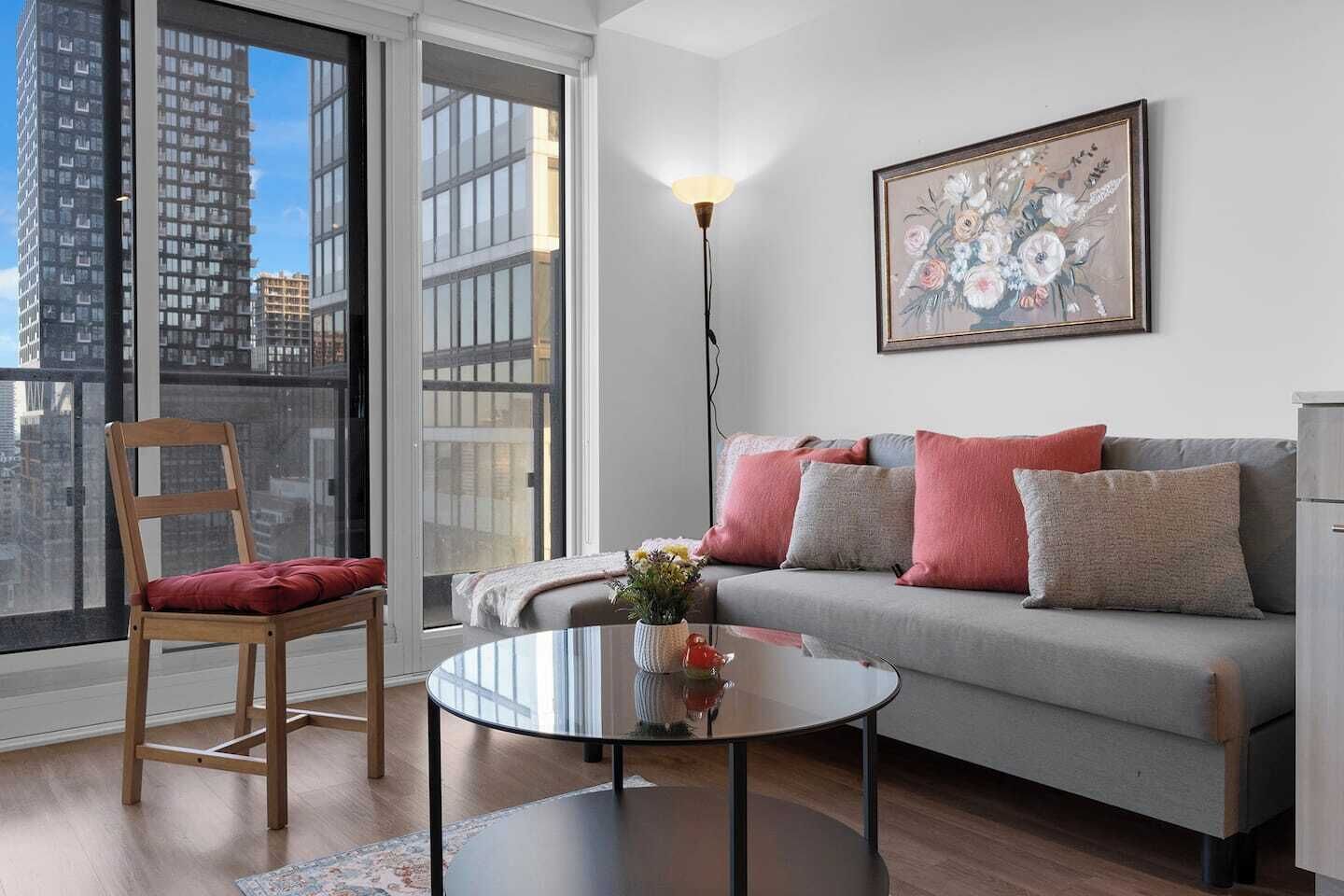 Ein modernes Wohnzimmer mit grauem Sofa, einem Holzstuhl, einem Glastisch und Blick auf die Stadt durch große Fenster.