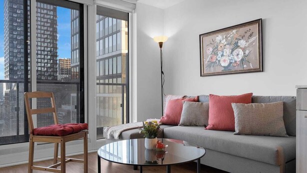 Ein modernes Wohnzimmer mit grauem Sofa, einem Holzstuhl, einem Glastisch und Blick auf die Stadt durch große Fenster.