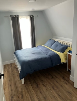 A bedroom with a bed, a nightstand, a window with curtains, and a wooden floor.