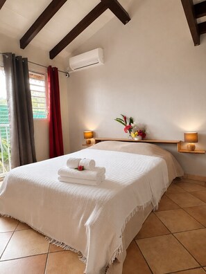 A hotel room with a large bed, two bedside lamps, a window with curtains, and an air conditioning unit. - Family Duplex 150 Meters from the Ocean (Les Trois-Îlets)