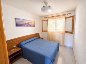 Et soveværelse med seng, natbord, ventilator i loftet, et vindue med gardiner og et billedværk på væggen.