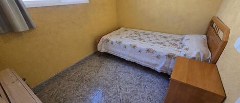 A small room with a single bed, a wooden nightstand, and a window with curtains.