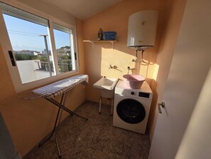 A compact laundry room with a washing machine, a sink, and an ironing board near a window. - Penisol 12 · 4 bedroom villa with pool (Peñíscola)