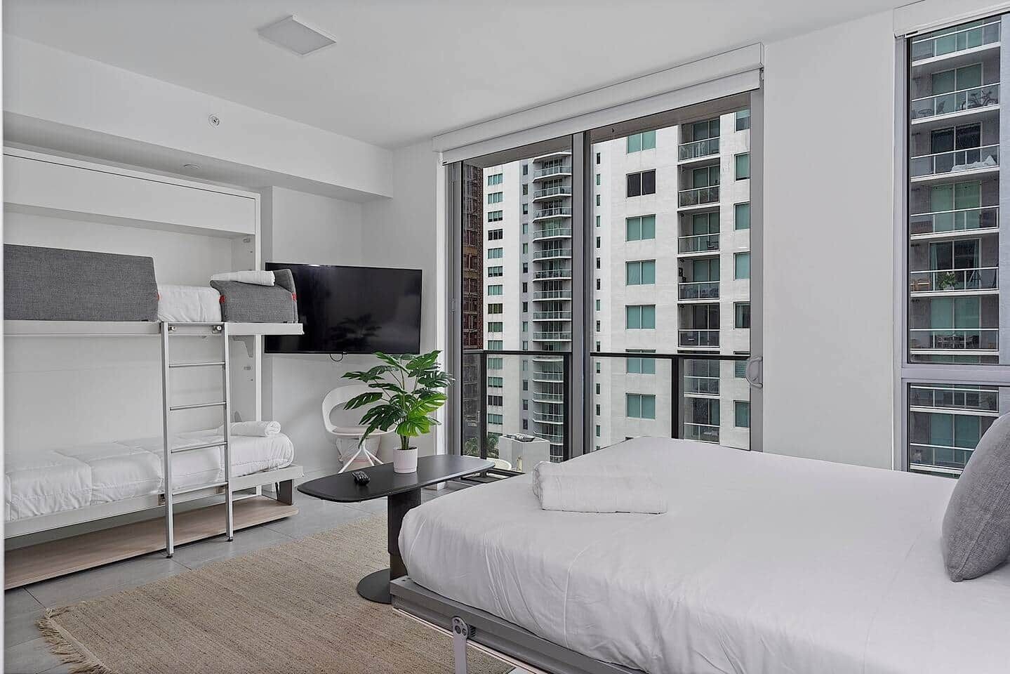A modern bedroom with a bed, a TV, a plant, and a view of a high-rise building.